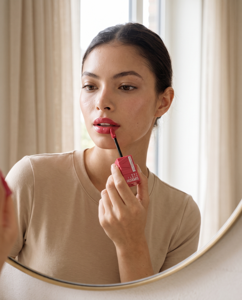 Model vor einem Spiegel beim Auftragen des Maybelline Superstay Vinyl Ink Lippenstifts in Pink